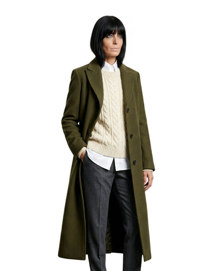 Person wearing a long olive green coat over a cream sweater and dark pants on a white background