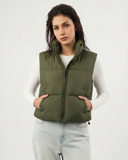 Woman wearing an olive green puffer vest over a white long-sleeve shirt on a light gray background