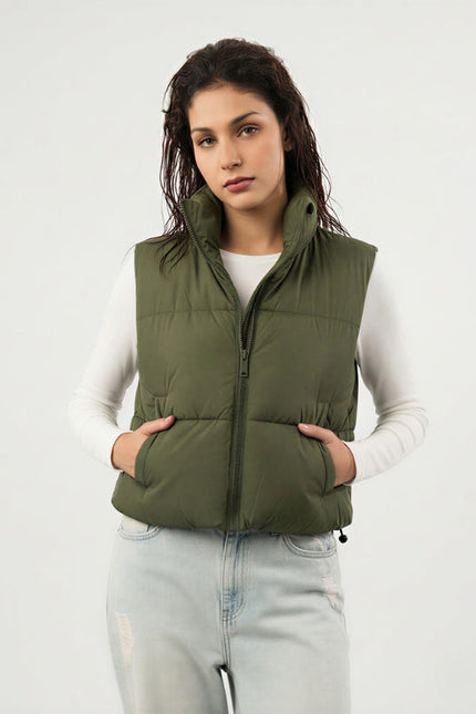 Woman wearing an olive green puffer vest over a white long-sleeve shirt on a light gray background