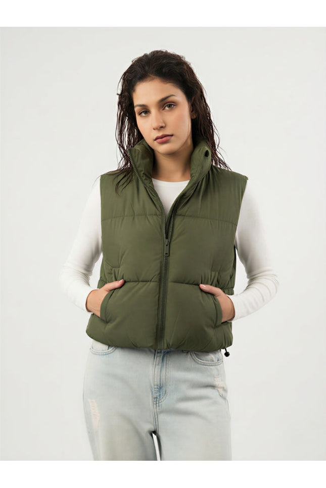 Woman wearing an olive green puffer vest over a white long-sleeve shirt on a light gray background