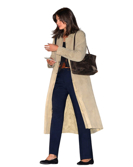 Woman in a beige trench coat and navy pants holding a phone and a brown handbag on a white background