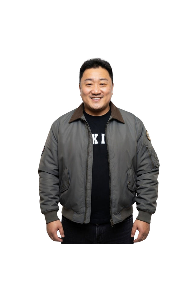 Man wearing a gray bomber jacket over a black shirt with visible branding on a white background