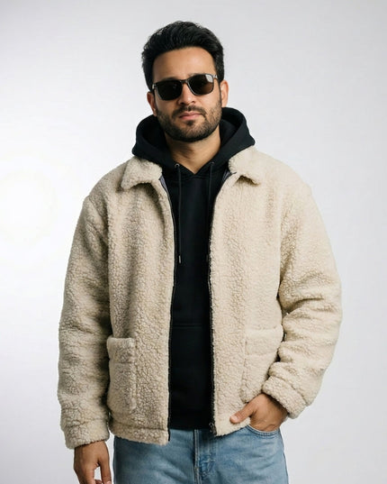 Man wearing a beige sherpa jacket, black hoodie, sunglasses, and blue jeans on a light gray background