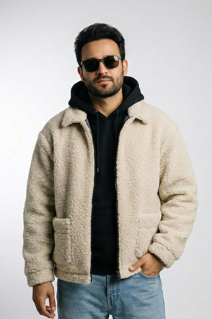 Man wearing a beige sherpa jacket, black hoodie, sunglasses, and blue jeans on a light gray background