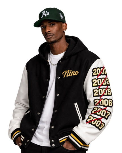 Man wearing a black and white letterman jacket with 'Nine' and years printed on sleeves, paired with a green baseball cap.