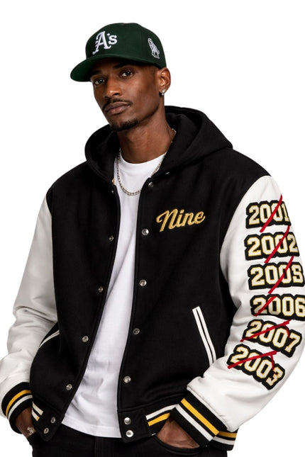 Man wearing a black and white letterman jacket with 'Nine' and years printed on sleeves, paired with a green baseball cap.