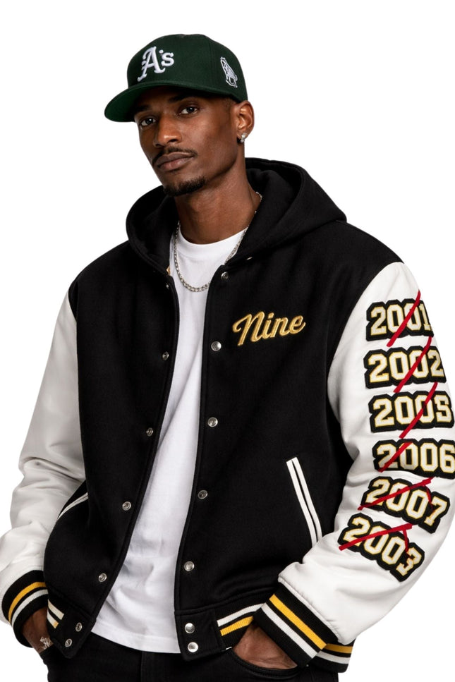 Man wearing a black and white letterman jacket with 'Nine' and years printed on sleeves, paired with a green baseball cap.