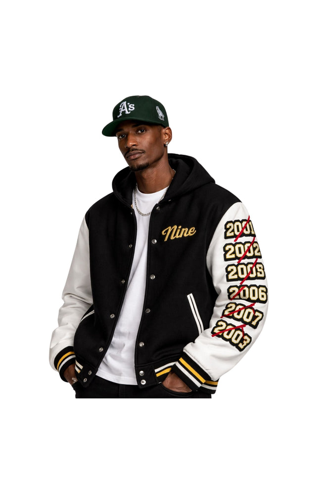 Man wearing a black and white letterman jacket with 'Nine' and years printed on sleeves, paired with a green baseball cap.