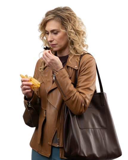 Woman in a brown leather jacket holding a brown leather handbag and eating a snack.