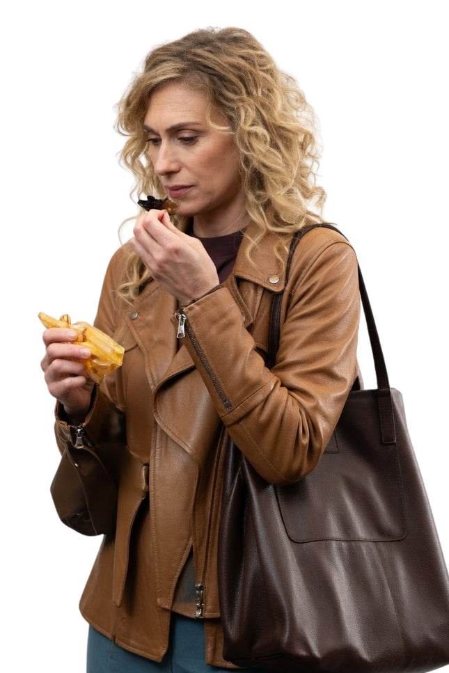 Woman in a brown leather jacket holding a brown leather handbag and eating a snack.