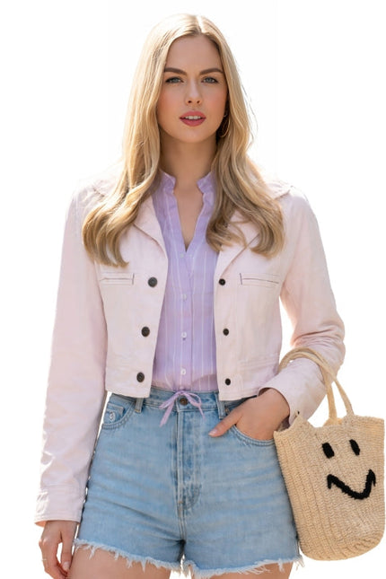 Woman wearing a light pink jacket, purple shirt, and denim shorts, holding a straw bag with a smiley face design.